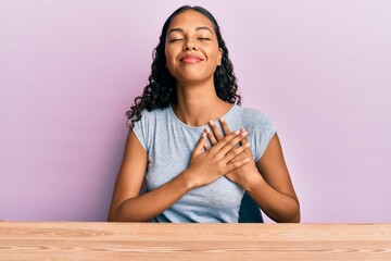 Young african american girl wearing casual clothes sitting on the table smiling with hands on chest with closed eyes and grateful gesture on face. health concept.