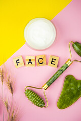 Green Gua Sha Facial Massage Tools. Roller made of green quartz jade on a pink-yellow background. Jar of cream, inscription face in wooden letters. Close-up, top view.
