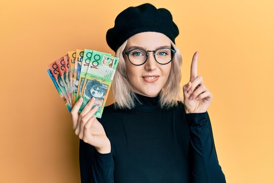 Young blonde girl holding australian dollars smiling with an idea or question pointing finger with happy face, number one