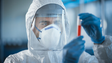 Medical Research Scientist Wearing Coverall, Surgical Gloves, Face Mask and Shield Holds Test Tube with Blood Sample and Label Reading Coronavirus. Microbiology Laboratory Drug and Vaccine Development