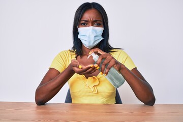 Young african american woman wearing medical mask holding hand sanitizer gel skeptic and nervous,...