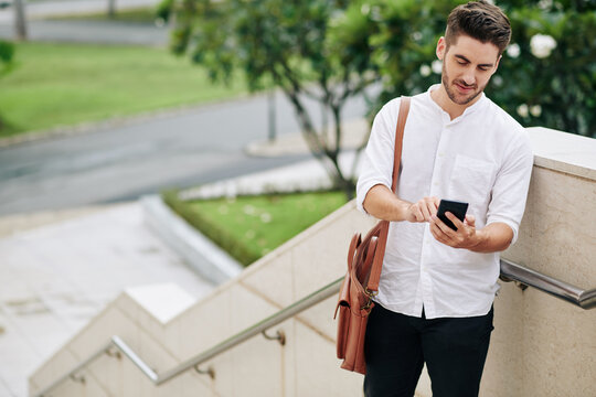 Handsome young man in white shirt standing on stairs and answering text message from friend or colleague
