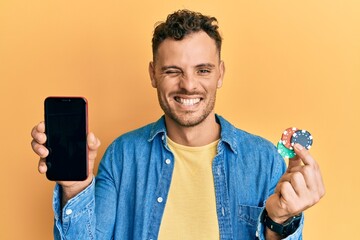 Young hispanic man holding poker chips and smartphone showing blank screen winking looking at the...