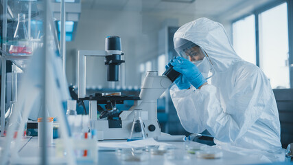 Beautiful Female Medical Scientist Wearing Coverall and Face Mask Looking at Petri Dish Under...
