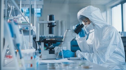 Beautiful Female Medical Scientist Wearing Coverall and Face Mask Looking at Petri Dish Under...