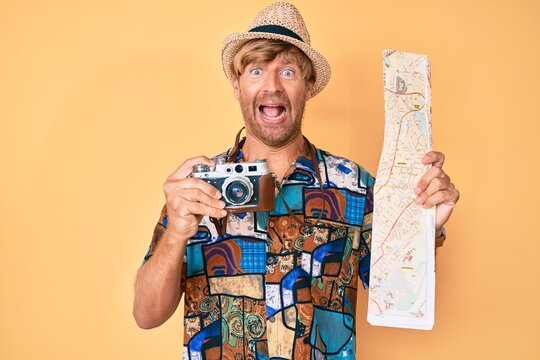 Young blond man holding vintage camera and city map celebrating crazy and amazed for success with open eyes screaming excited.