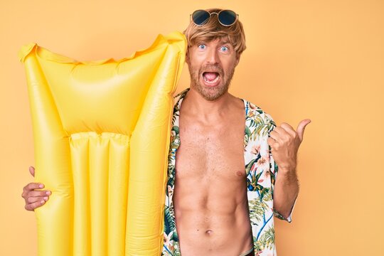 Young blond man wearing summer style and holding matress float pointing thumb up to the side smiling happy with open mouth