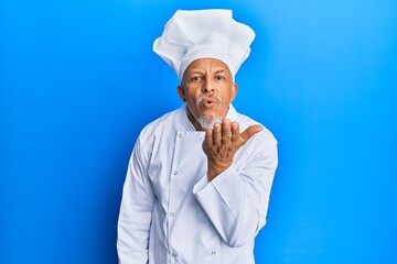 Middle age grey-haired man wearing professional cook uniform and hat looking at the camera blowing a kiss with hand on air being lovely and sexy. love expression.