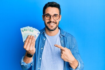 Young hispanic man holding bunch of 20 euro banknotes smiling happy pointing with hand and finger
