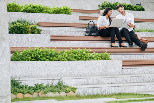 Cheerful young business people sitting on bench with laptop and discussing marketing strategy and new project developement