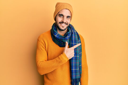Young hispanic man wearing wool sweater and winter scarf smiling cheerful pointing with hand and finger up to the side