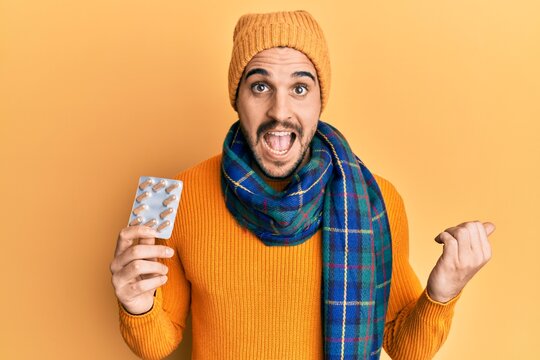 Young hispanic man wearing wool sweater and winter scarf holding pills pointing thumb up to the side smiling happy with open mouth