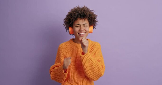 Carefree teenage girl listens favorite music keeps hand like microphone sings along moves with every bit wears stereo headphones orange casual jumper isolated over purple background. Real meloman