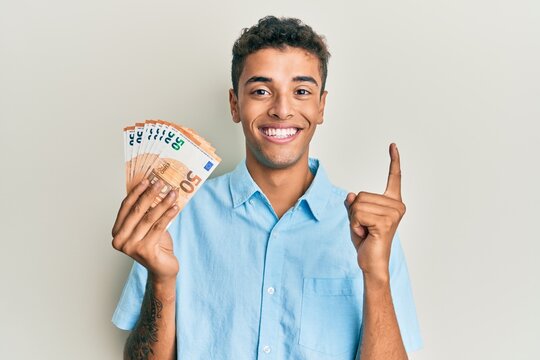 Young handsome african american man holding bunch of 50 euro banknotes smiling with an idea or question pointing finger with happy face, number one