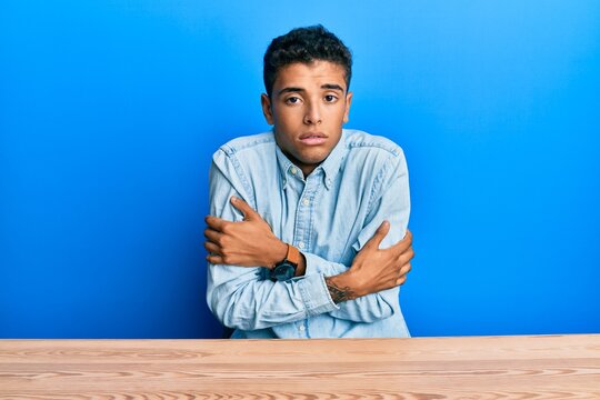 Young handsome african american man wearing casual clothes sitting on the table shaking and freezing for winter cold with sad and shock expression on face