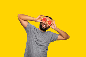 portrait of a man in gray t-shirt has fun with grapefruit. guy closes his eyes with fruit, smile. yellow background. isolated 