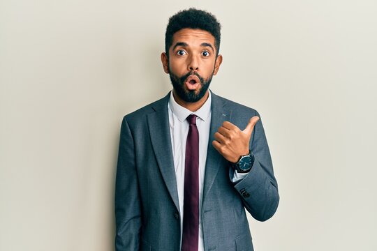 Handsome hispanic man with beard wearing business suit and tie surprised pointing with hand finger to the side, open mouth amazed expression.