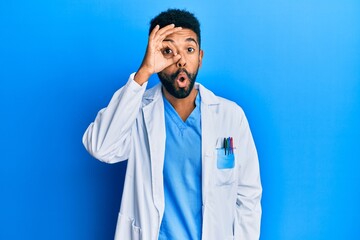 Handsome hispanic man with beard wearing doctor uniform doing ok gesture shocked with surprised face, eye looking through fingers. unbelieving expression.