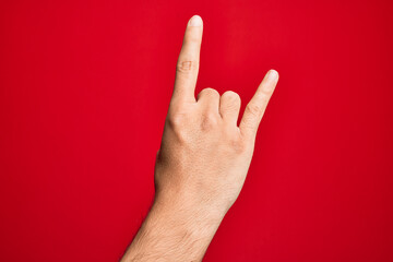 Hand of caucasian young man showing fingers over isolated red background gesturing rock and roll symbol, showing obscene horns gesture