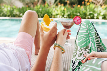 Obraz premium Cropped image of couple drinking cocktails when relaxing on chaise-lounges by swimming pool