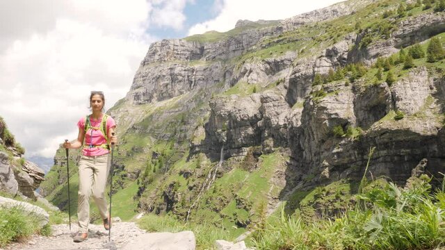 A gorgeous young woman in a pink t-shirt and hiking gear is walking towards the camera with a majestic mountain-scape in the background, slow motion.
