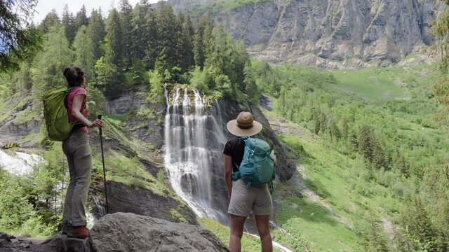Two young gorgeous hikers approach a grandiose waterfall and watch it in awe before exchanging a high five during the hike to the refuge de Salles, sixt-fer-à-cheval