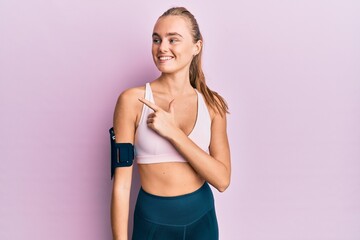 Beautiful blonde woman wearing sportswear and arm band cheerful with a smile of face pointing with hand and finger up to the side with happy and natural expression on face