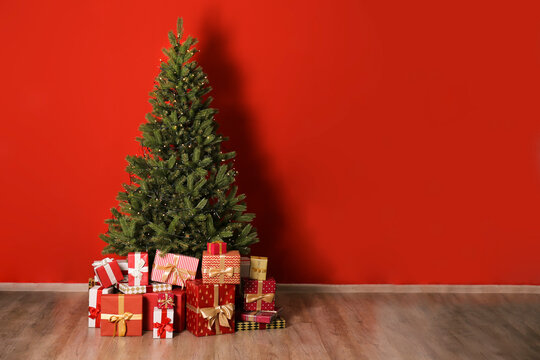 Big beautiful christmas tree decorated with just lights in minimal style and many different presents on wooden floor. Red wall background with a lot of copy space for text. Close up.