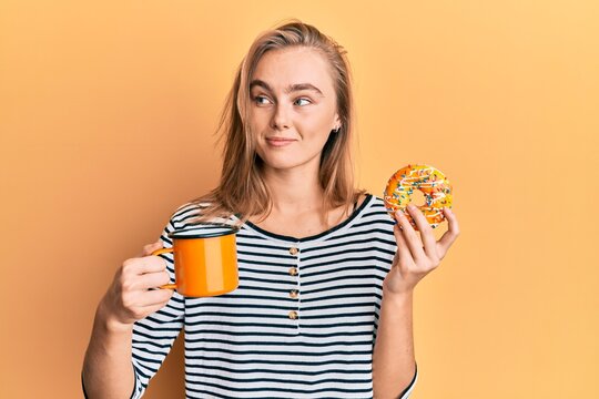 Beautiful blonde woman eating doughnut and drinking coffee smiling looking to the side and staring away thinking.