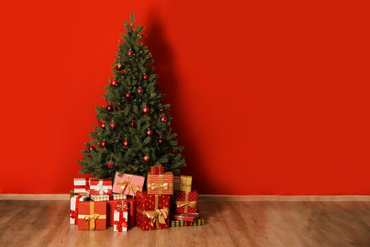 Big beautiful christmas tree decorated with beautiful shiny baubles in minimal style and many different presents on wooden floor. Red wall background with a lot of copy space for text. Close up.