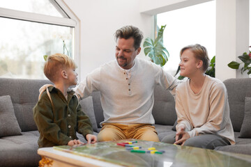 Obraz premium Portrait of happy father embracing two sons while playing board games together at home, copy space