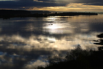 Fototapeta premium Night river.The black sky with Golden stripes is reflected in the water with ripples on the surface.In the center of the water area is a distant silhouette of a fisherman in a boat in the moonlight