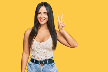 Young beautiful hispanic girl wearing casual clothes smiling with happy face winking at the camera doing victory sign. number two.