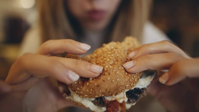 Macro close up of Young woman eating burger in fast food restaurant. People eat unhealthy meal. American style. Getting satisfaction from unhealthy food. Junk food, modern life concet.