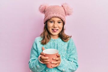 Little beautiful girl holding pink mug wearing cute wool hat sticking tongue out happy with funny expression.