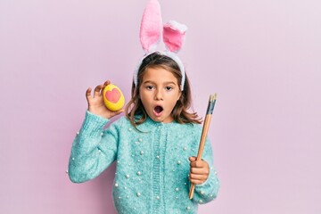 Little beautiful girl wearing cute easter bunny ears holding colored egg afraid and shocked with...
