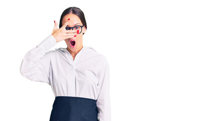 Beautiful brunette young woman wearing professional waitress apron peeking in shock covering face and eyes with hand, looking through fingers with embarrassed expression.