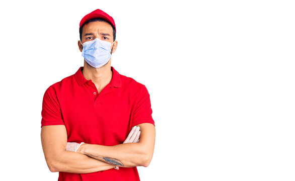 Young handsome hispanic man wearing delivery uniform and medical mask skeptic and nervous, disapproving expression on face with crossed arms. negative person.
