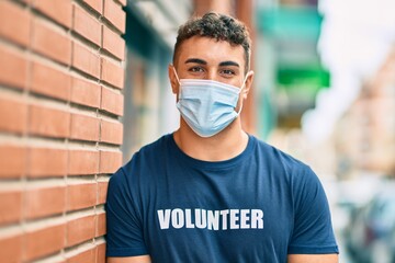 Young hispanic volunteer man wearing medical mask standing at the city.