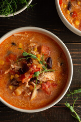 Spicy mexican soup with beans and corn, tomatoes, chicken breast and bean sprouts, bowl with soup and napkin on a dark wooden background, top view
