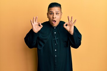 Young latin priest man standing over yellow background looking surprised and shocked doing ok...