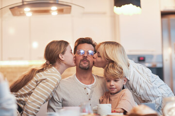 Portrait of happy father enjoying kisses from three children while sitting at dining table in kitchen
