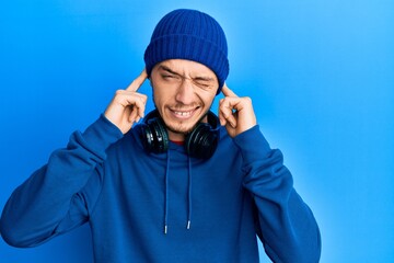 Hispanic young man wearing sweatshirt and headphones covering ears with fingers with annoyed expression for the noise of loud music. deaf concept.