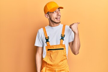 Hispanic young man wearing handyman uniform smiling with happy face looking and pointing to the side with thumb up.