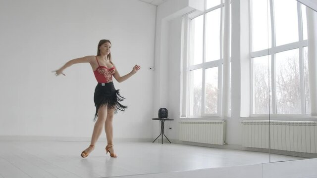Attractive girl dances ballroom dancing in front of a mirror in a bright white hall. A professional ballroom dancer trains in a choreographic hall.