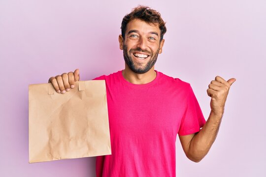 Handsome man with beard holding take away paper bag pointing thumb up to the side smiling happy with open mouth