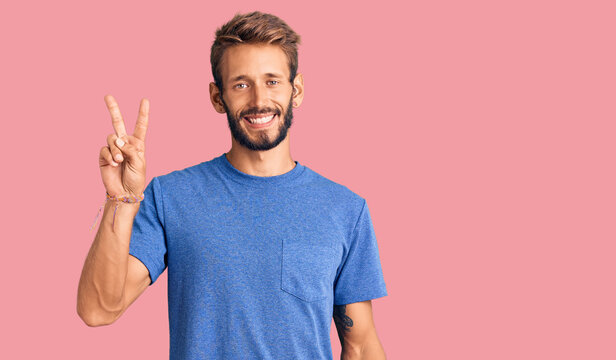 Handsome blond man with beard wearing casual clothes smiling with happy face winking at the camera doing victory sign. number two.