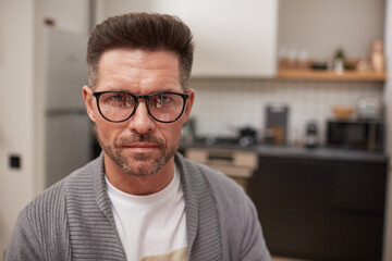 Fototapeta na wymiar Portrait of handsome mature man wearing glasses and looking at camera with serious face expression while sitting in kitchen interior at home, copy space