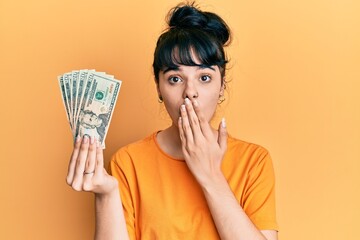 Young hispanic girl holding dollars covering mouth with hand, shocked and afraid for mistake....