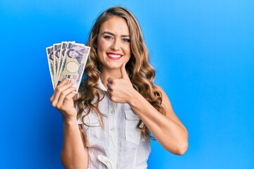 Young blonde girl holding japanese yen banknotes smiling happy and positive, thumb up doing excellent and approval sign